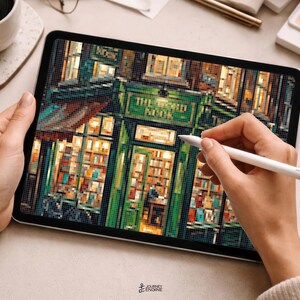 May include: A digital tablet displaying a pixel art illustration of a green storefront with the text THE BOOK NOOK visible above the entrance. The artwork features warm interior lighting, shelves filled with books, and a detailed facade design.