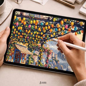 May include: A tablet displaying a pixelated digital art piece of a coastal town with colorful hanging lanterns, vibrant buildings, and greenery. A white stylus is held against the screen, which features the small text Journey Engine at the bottom.