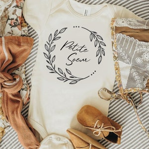 Peut inclure: Body de bébé crème avec l'inscription "Petite Soeur" dans une couronne florale noire. L'image comprend des chaussures de bébé marron, des chaussettes et un bonnet fleuri. Le texte en haut indique "DISPONIBLE ÉGALEMENT EN D'AUTRES COLORIS EN MANCHES COURTES OU LONGUES".