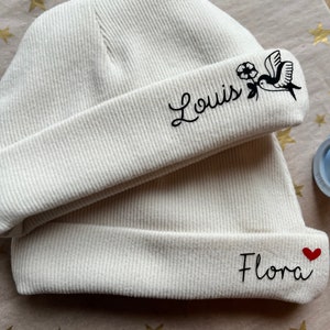 May include: Two white knitted beanies with embroidered names. The beanie on top has a small bird and flower design and the name "Louis" embroidered on it. The beanie on the bottom has a red heart and the name "Flora" embroidered on it.