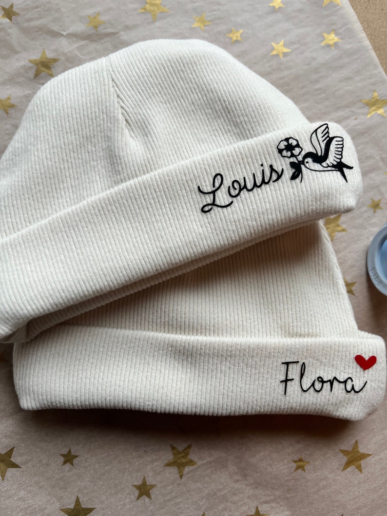 May include: Two white knitted beanies with embroidered names. The beanie on top has a small bird and flower design and the name "Louis" embroidered on it. The beanie on the bottom has a red heart and the name "Flora" embroidered on it.