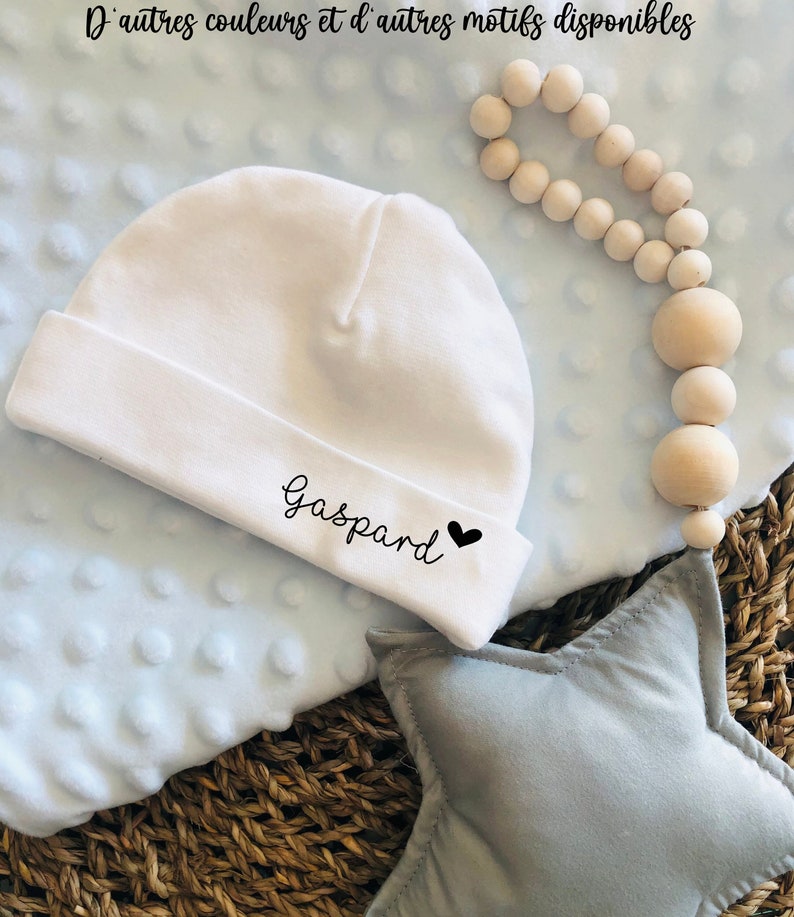 May include: White baby hat with the name "Gaspard" and a black heart embroidered on it. The hat is on a blue blanket with a star-shaped pillow.