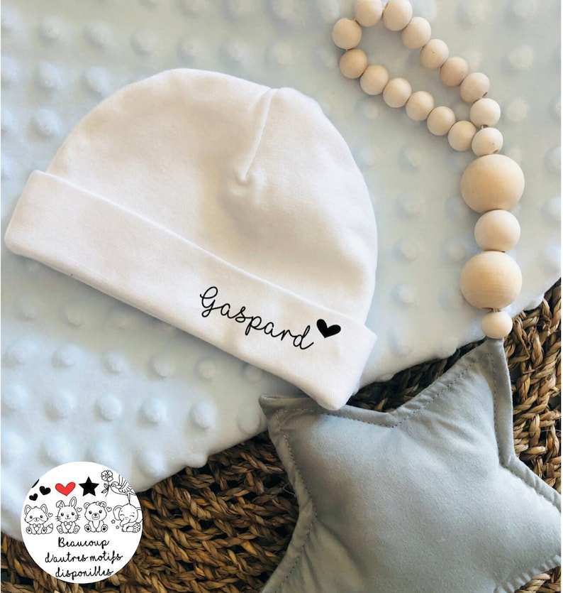 May include: White baby hat with the name "Gaspard" and a small black heart embroidered on the front. The hat is on a blue blanket with a star-shaped pillow and a wooden bead necklace.