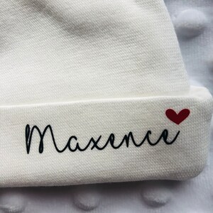 May include: White baby hat with the name "Maxence" embroidered in black cursive lettering and a small red heart.
