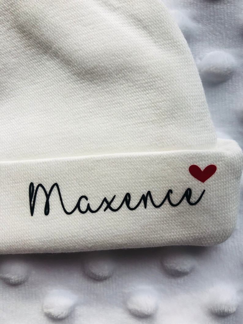May include: White baby hat with the name "Maxence" embroidered in black cursive lettering and a small red heart.