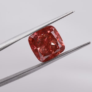 Op de afbeelding: Een vierkant geslepen, roodachtig-oranje diamant, vastgehouden door een zilveren pincet tegen een witte achtergrond. De edelsteen heeft een gefacetteerd oppervlak dat licht reflecteert. De pincet staat diagonaal in het frame.