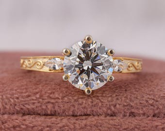 May include: A gold ring featuring a large round brilliant-cut center stone held by six prongs. The band is decorated with engraved scrollwork patterns and includes a small marquise-cut stone on each side of the central setting.