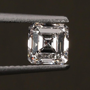 May include: A square-cut, clear diamond held by tweezers against a dark background. The diamond's facets reflect light, creating a brilliant sparkle. The gem is a classic shape, ideal for jewelry.
