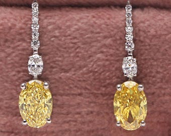 May include: A pair of elegant drop earrings featuring a vibrant yellow oval-cut gemstone. The earrings have a delicate design with a row of small, sparkling white stones and a larger white stone above the yellow gemstone. The earrings are set in a silver metal.