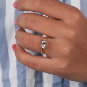 以下が含まれることがあります： マーキスカットのダイヤモンドをあしらったゴールドリング。ダイヤモンドは繊細なバンドにセットされています。ピンクのマニキュアを塗った手の上に、青と白のストライプの背景にリングが写っています。