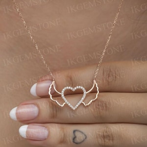 Puede incluir: Collar de oro rosa con un colgante en forma de corazón adornado con diamantes y alas. La fina y elegante cadena se muestra en una mano. Una hermosa pieza de joyería, perfecta para regalar.