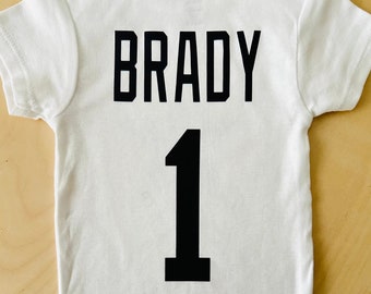 May include: White baby onesie with black text that says "BRADY" and the number "1" on the back.
