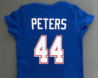 Puede incluir: Un body de bebé azul real con el nombre "PETERS" impreso en letras blancas en la parte superior de la espalda. Debajo del nombre, el número "44" se muestra en blanco con un contorno rojo. El body es de manga corta.