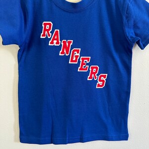 May include: A blue short-sleeved t-shirt with the word "RANGERS" in red letters on the front. The shirt has a white tag that says "Gerber Kids" on it.