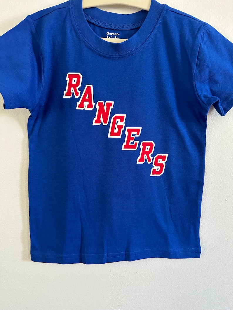 May include: A blue short-sleeved t-shirt with the word "RANGERS" in red letters on the front. The shirt has a white tag that says "Gerber Kids" on it.
