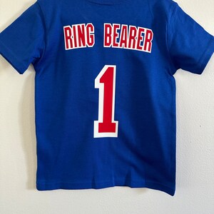 May include: A blue t-shirt with the words "Ring Bearer" and the number "1" in red and white lettering on the back.