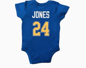 Può includere: Un body blu per neonato con il nome "JONES" e il numero "24" stampato sul retro in giallo.