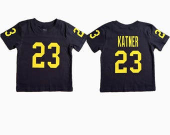 Könnte beinhalten: Zwei dunkelblaue T-Shirts mit kurzen Ärmeln. Die Vorderseite des ersten T-Shirts zeigt die gelbe Nummer 23 in der Mitte sowie die Nummer 3 auf dem rechten Ärmel und die 2 auf dem linken. Das zweite T-Shirt zeigt den Namen KATNER und die Nummer 23.