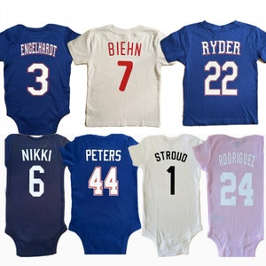 May include: Six baby onesies with different names and numbers printed on the back. The onesies are in blue, white, and pink.