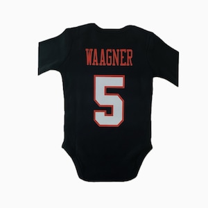 Op de afbeelding: Zwarte babybody met lange mouwen. De achterkant toont de naam "WAAGNER" in rode blokletters boven het nummer "5" in wit met een rode omtrek. De body is ontworpen voor baby's.