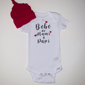 May include: White baby onesie with black text that reads "Bebé de Mami y Papi" with red hearts. A red knit baby hat is included.
