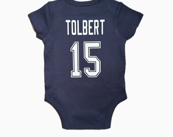 Pode incluir: Um macacão de bebê azul escuro com texto branco que diz "TOLBERT 15".