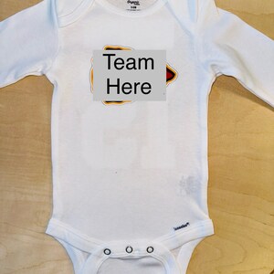 Op de afbeelding: Witte baby romper met lange mouwen met een rood, geel en zwart strijkmotief met de tekst "Team Here".