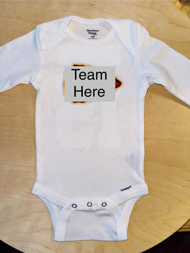 Op de afbeelding: Witte baby romper met lange mouwen met een rood, geel en zwart strijkmotief met de tekst "Team Here".