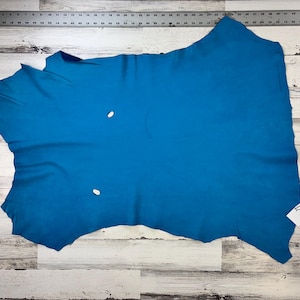 Puede incluir: Un trozo de cuero azul turquesa con una forma ligeramente irregular. El cuero está colocado plano sobre una superficie de madera blanca. El cuero tiene dos pequeños agujeros.