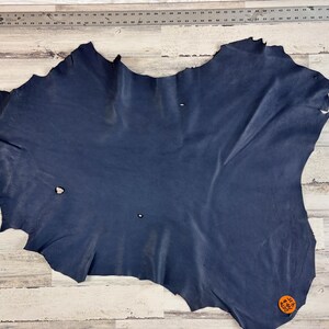 May include: A large, irregularly shaped piece of navy blue leather. The leather has a textured surface with natural variations in color and tone. A metal ruler is visible along the top edge, and a small orange tag is attached to the leather.