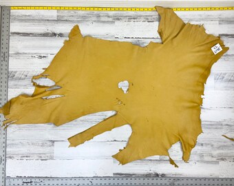May include: A large piece of yellow leather with some holes and tears. The leather is laid out on a white wooden surface. A tag with the text "S.L. 50-50" is attached to the leather.