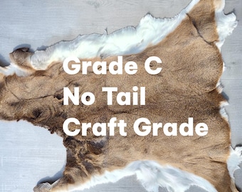 May include: A deer hide with brown and white fur, labeled "Grade C No Tail Craft Grade". The hide is laid out on a light gray surface. The fur has a natural, textured appearance.