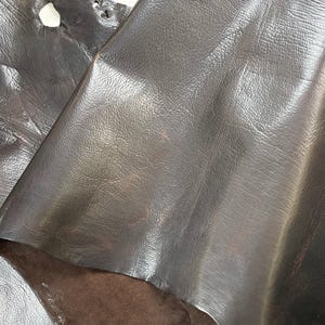 Puede incluir: Primer plano de hojas de cuero marr&oacute;n oscuro. El cuero tiene una superficie lisa y ligeramente brillante con sutiles variaciones de color y textura. La imagen muestra la veta natural y los pliegues del material, destacando su calidad flexible. El cuero parece ser de alta calidad.