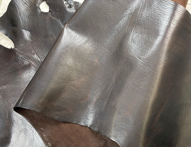 Puede incluir: Primer plano de hojas de cuero marr&oacute;n oscuro. El cuero tiene una superficie lisa y ligeramente brillante con sutiles variaciones de color y textura. La imagen muestra la veta natural y los pliegues del material, destacando su calidad flexible. El cuero parece ser de alta calidad.