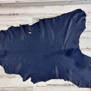 May include: A large, irregularly shaped piece of navy blue leather. The leather has a textured surface and varying shades of blue. A metal ruler with inch measurements is visible along the left edge. The leather is lying on a light-colored wooden surface.