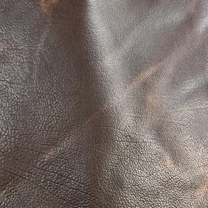 Puede incluir: Primer plano de cuero marr&oacute;n oscuro con una superficie texturizada. El cuero tiene un aspecto envejecido con variaciones de color, mostrando &aacute;reas marrones m&aacute;s claras y pliegues sutiles. La imagen resalta la veta natural y las imperfecciones del material.