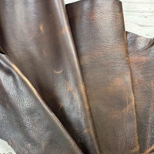 Puede incluir: Primer plano de varias piezas de cuero marrón oscuro con un aspecto envejecido y texturizado. El cuero presenta variaciones de color, con zonas marrón claro y grano visible. Los pliegues y arrugas añaden profundidad y carácter al material.