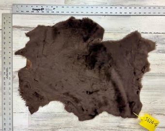 Może przedstawiać: Ciemnobrązowa skóra owcza o miękkiej, teksturowanej powierzchni. Skóra ma nieregularny kształt i ma około 76 cm długości. Dołączona jest żółta etykieta z napisem "S624-9".