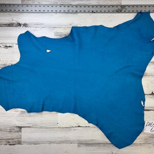 May include: A piece of turquoise blue leather with a small hole in the centre. The leather is laid out on a white wooden surface with a ruler and a tag that says "D164TQ0 62".
