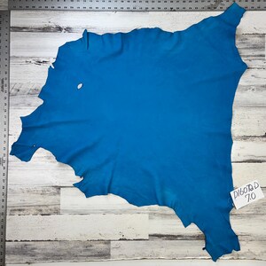 Puede incluir: Un trozo de cuero azul turquesa de forma irregular. El cuero está colocado sobre una superficie de madera blanca. Una pequeña etiqueta blanca con el texto "D1607D 7.0" está unida al cuero.