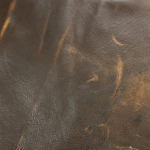 Puede incluir: Primer plano de cuero marr&oacute;n desgastado con reflejos marr&oacute;n claro. El cuero tiene una superficie texturizada con grano visible y sutiles variaciones de color, creando una apariencia vintage o r&uacute;stica. La imagen muestra las imperfecciones naturales y el car&aacute;cter del material.