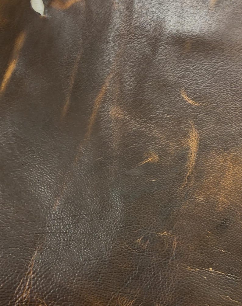 Puede incluir: Primer plano de cuero marr&oacute;n desgastado con reflejos marr&oacute;n claro. El cuero tiene una superficie texturizada con grano visible y sutiles variaciones de color, creando una apariencia vintage o r&uacute;stica. La imagen muestra las imperfecciones naturales y el car&aacute;cter del material.