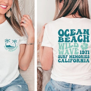 May include: White t-shirt with a front pocket design featuring palm trees, waves, and the text "Surf Memories." The back of the shirt displays the words "Ocean Beach Wild Wave 1971 Surf Memories California" in teal and blue.