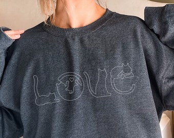 以下が含まれることがあります： 白い文字で「LOVE」と書かれた濃いグレーのスウェットシャツ。各文字は猫のシルエットでデザインされています。スウェットシャツは長袖とクルーネックです。デザインはシンプルでモダンです。