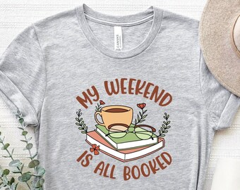 Może przedstawiać: Szara koszulka z napisem "My Weekend Is All Booked" w brązowych literach. Projekt zawiera filiżankę kawy, okulary i stos książek.
