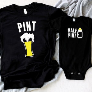 以下が含まれることがあります： ビールグラスと哺乳瓶のグラフィックが描かれた黒いTシャツ2枚。大きい方のTシャツには「PINT」、小さい方のTシャツには「HALF PINT」と書いてあります。