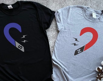 Puede incluir: Dos camisetas con un gráfico de un imán en forma de corazón. La camiseta de la izquierda es negra con un imán azul y la letra "S" en ella. La camiseta de la derecha es gris con un imán rojo y la letra "N" en ella.