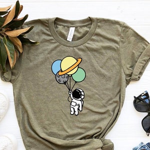 May include: A green t-shirt with a cartoon astronaut holding balloons shaped like planets. The planets are yellow, blue, green, and gray.