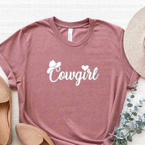 K&ouml;nnte beinhalten: Ein mauvefarbenes T-Shirt mit einer wei&szlig;en Grafik eines Cowboyhuts und dem Wort "Cowgirl" in kursiv.