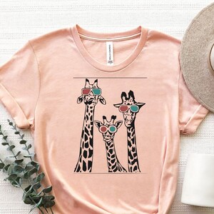 May include: A peach-coloured t-shirt featuring a graphic of three giraffes wearing 3D glasses. The giraffes are black and white with brown spots.